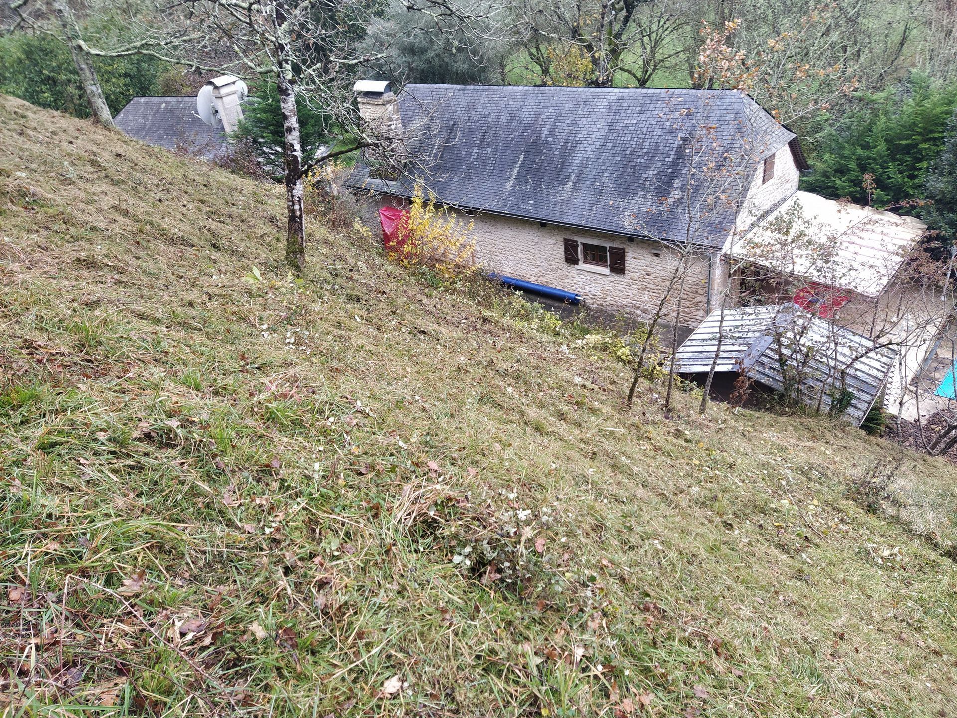 Maison en pierre avec toit en ardoise à flanc de colline, entourée d'arbres et d'herbe.