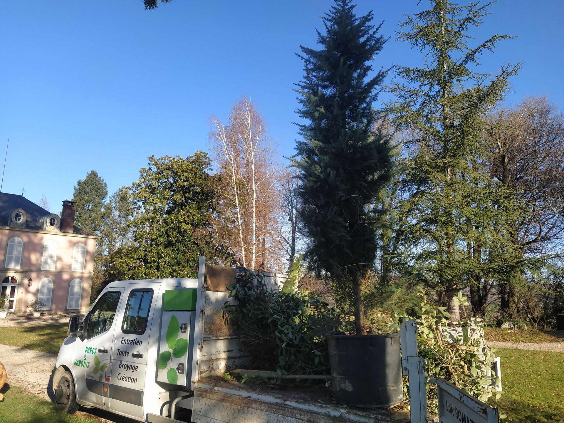 Une camionnette blanche garée près d'un bâtiment et d'arbres par une journée ensoleillée. Le texte est vert et blanc.