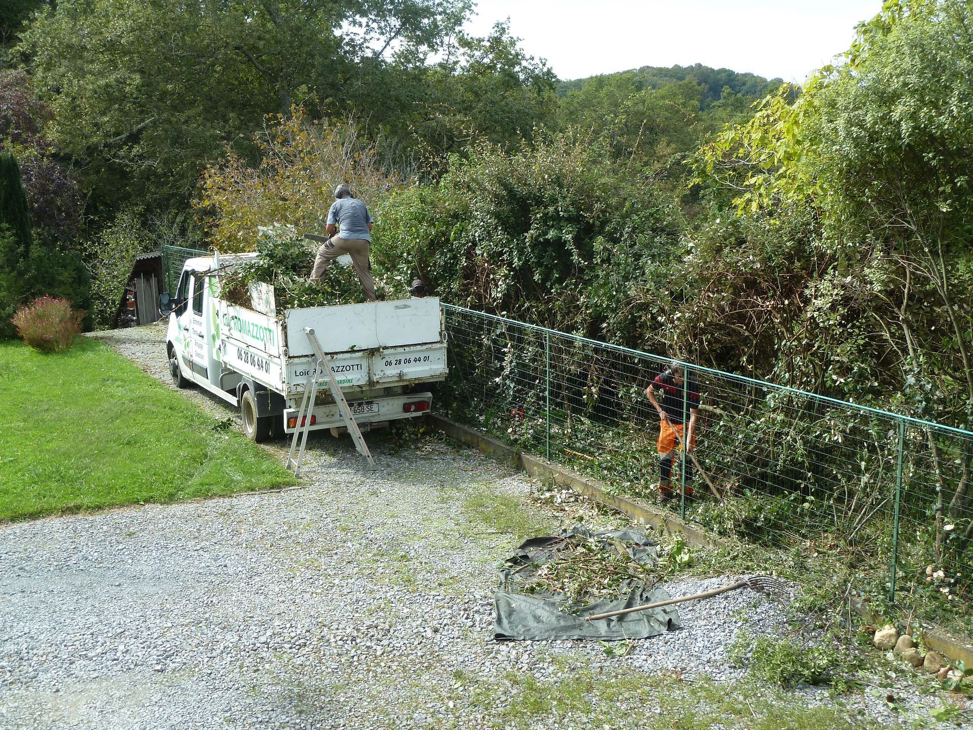 Des hommes taillent des buissons, chargent des débris dans un camion, à côté d'une allée de gravier et d'une pelouse verte.
