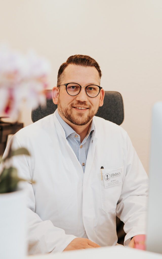Ein lächelnder Arzt im weißen Kittel und mit Brille sitzt in einem hellen Büro.