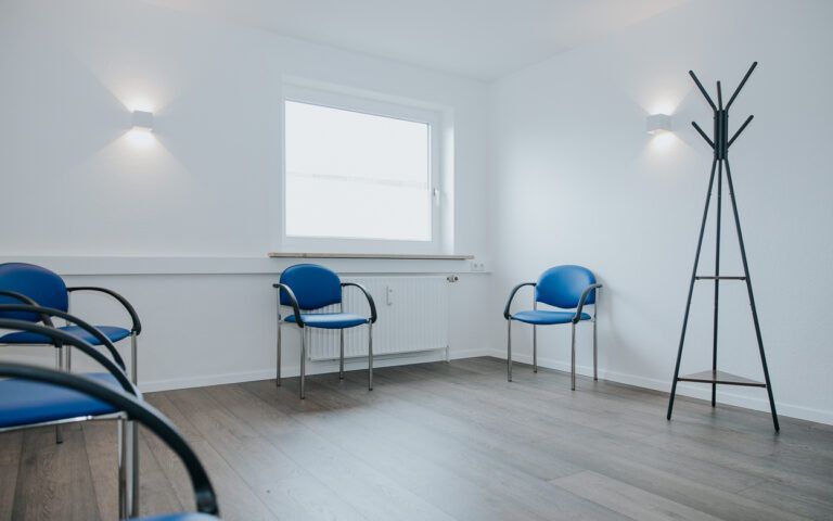 Leerer Warteraum mit blauen Stühlen, weißen Wänden, einem Fenster und einer schwarzen Garderobe