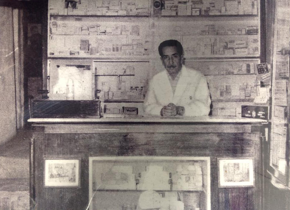 Foto storica con Dottor Messana, uomo in camice bianco dietro il bancone della farmacia. Dietro di lui, scaffali pieni di prodotti confezionati.