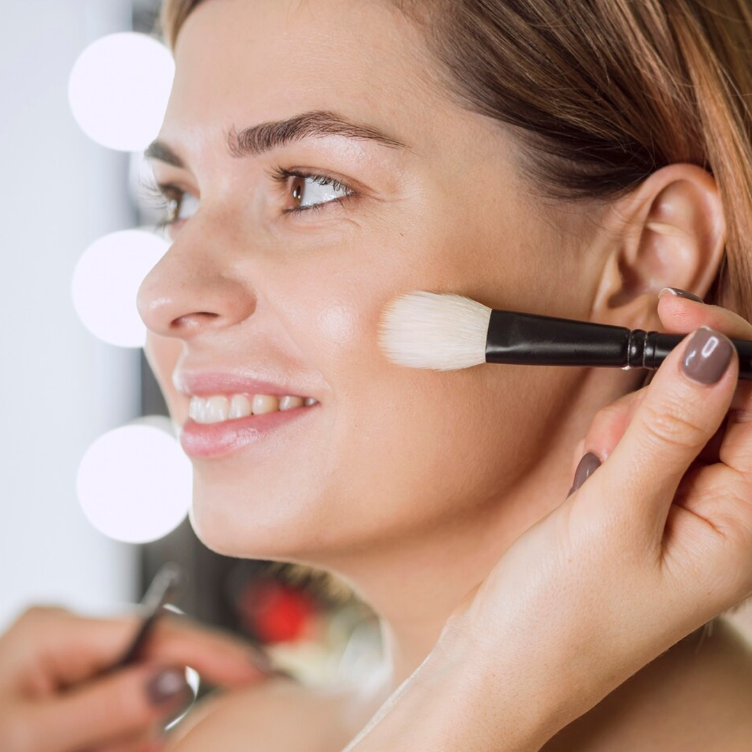 Persona sorridente mentre il trucco viene applicato con un pennello sulla guancia, illuminata dalle luci dello studio.
