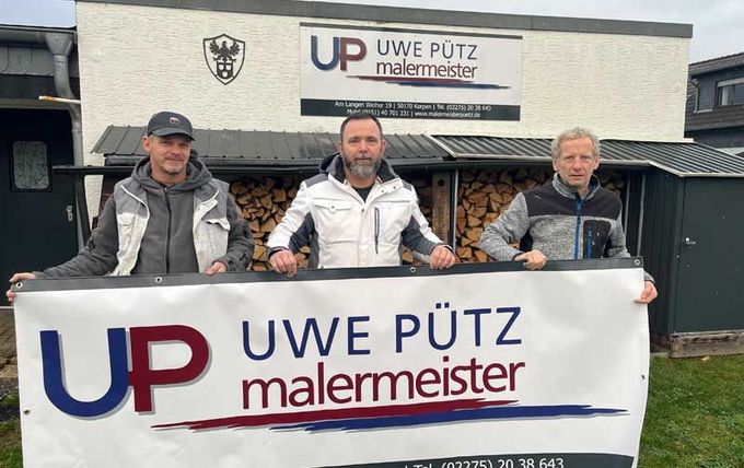 Drei Männer halten vor einem Gebäude ein Schild mit der Aufschrift „Uwe Putz Malermeister“.