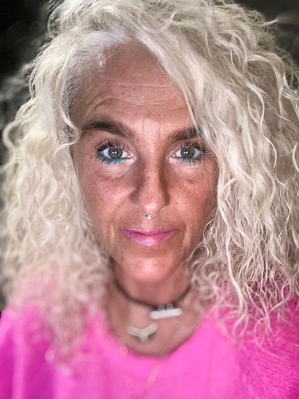 Eine Frau mit blonden Locken und blauen Augen trägt ein rosa Hemd.