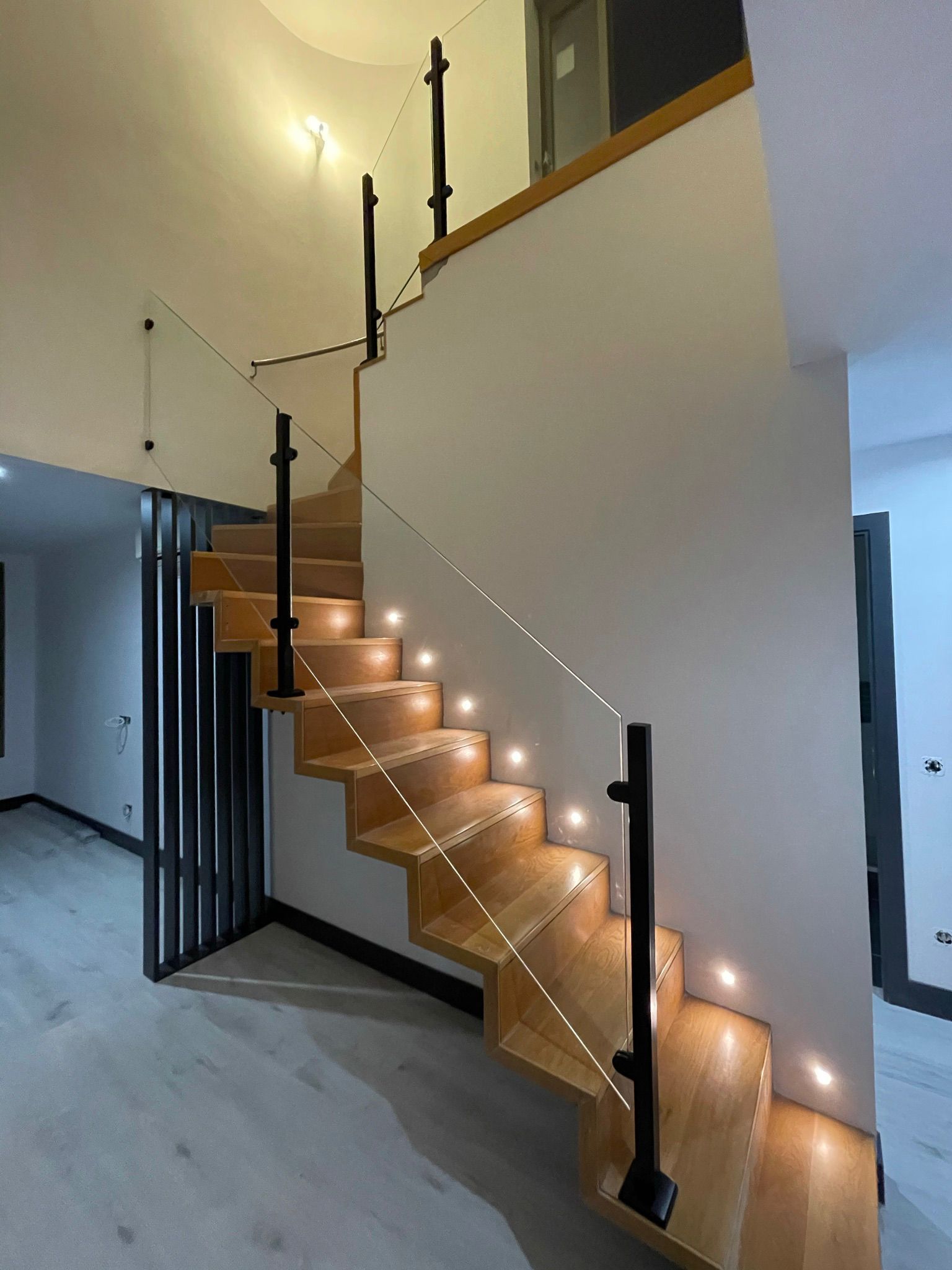 Escalera de madera con luces integradas, barandillas negras y panel de vidrio contra una pared blanca.