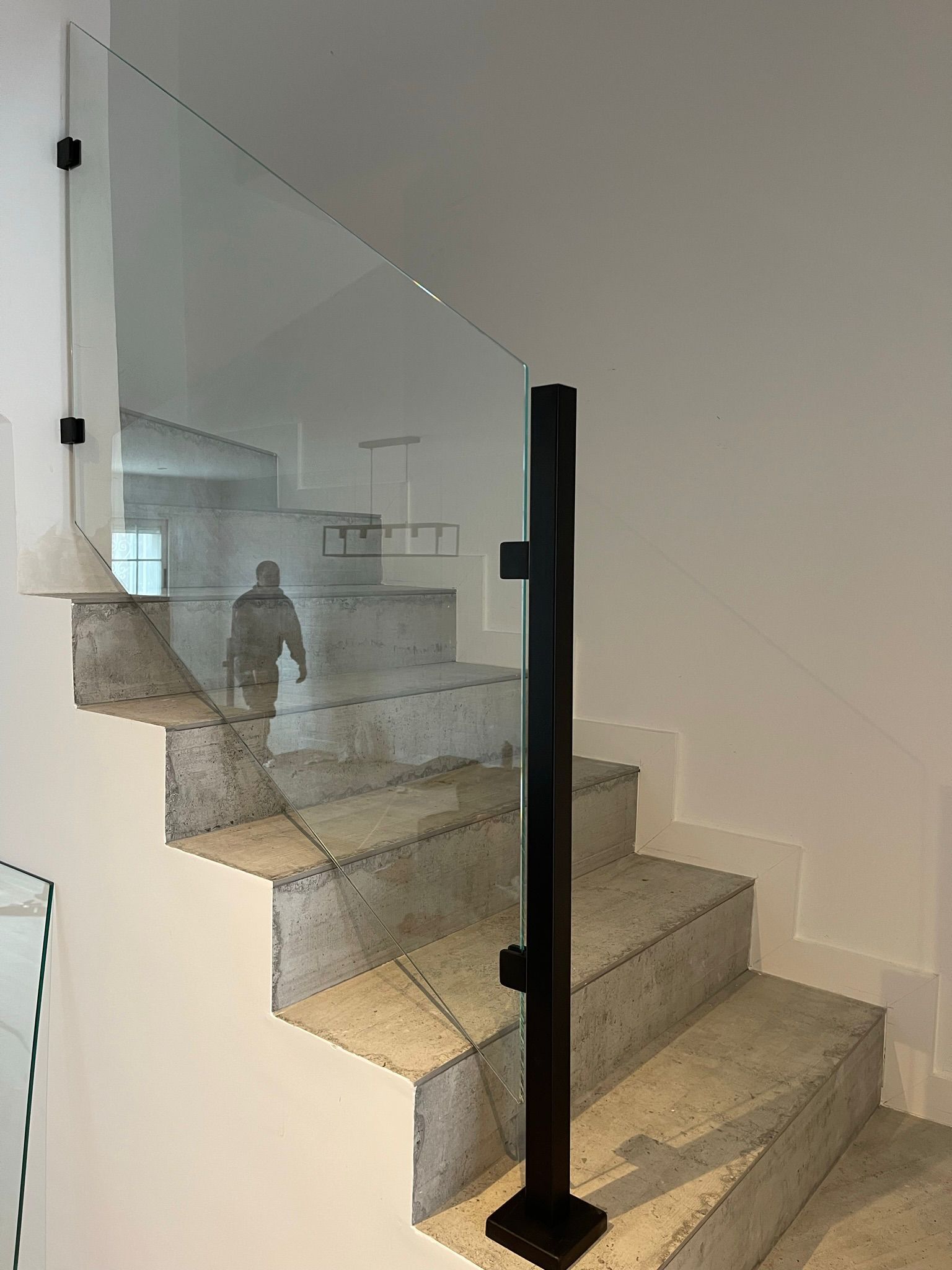 Escalera con escalones de hormigón, barandilla de cristal y postes de soporte negros. Se ve una persona reflejada.