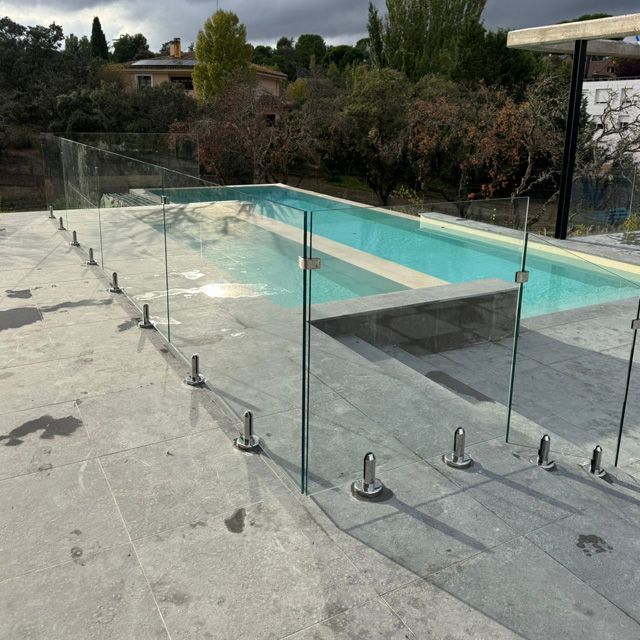 Piscina con valla de cristal transparente, situada en un patio de piedra gris, rodeada de árboles y un cielo nublado.