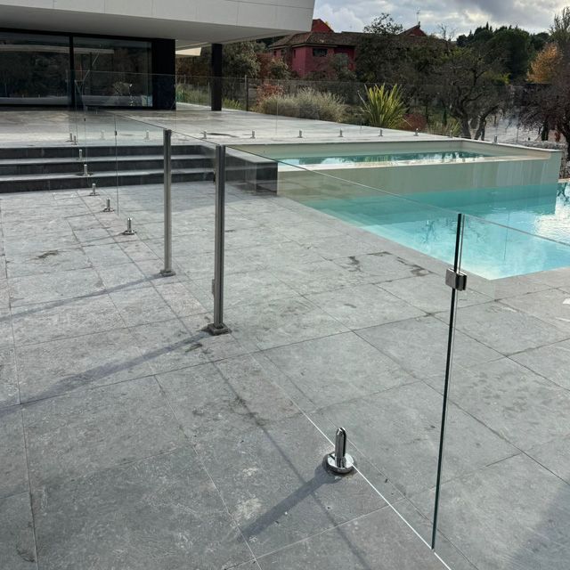 Valla de cristal para piscina en un patio de piedra, con una piscina transparente y una casa moderna al fondo.