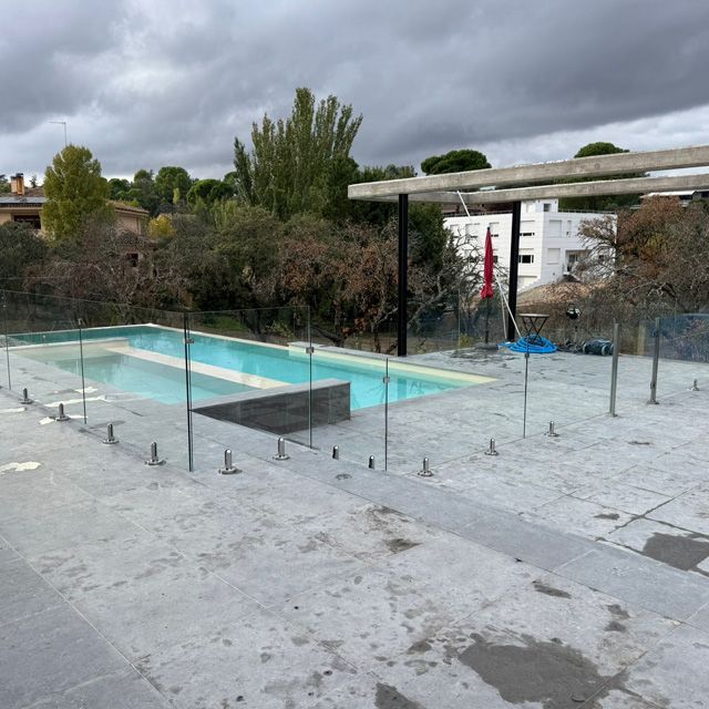 Piscina rodeada de valla de cristal y patio gris bajo un cielo nublado.