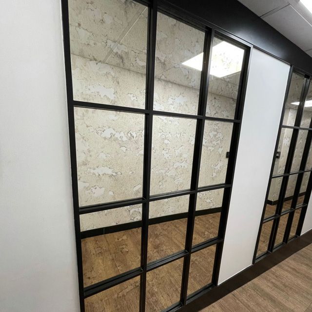 Paneles de ventana con marco negro que reflejan una habitación con pared texturizada, piso de madera y paredes blancas.