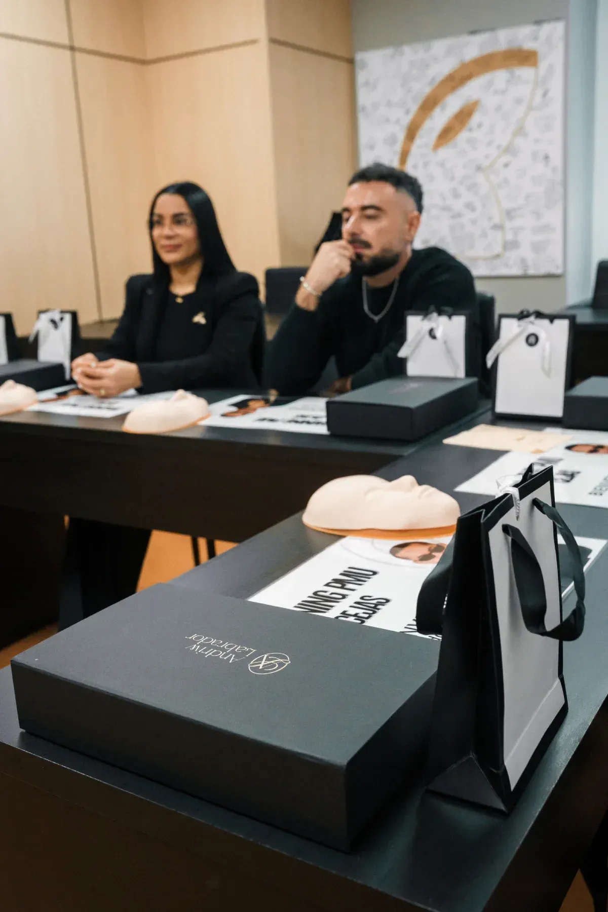 Dos personas sentadas a una mesa con cajas de regalo negras y bolsas de la compra blancas en una sala de reuniones.