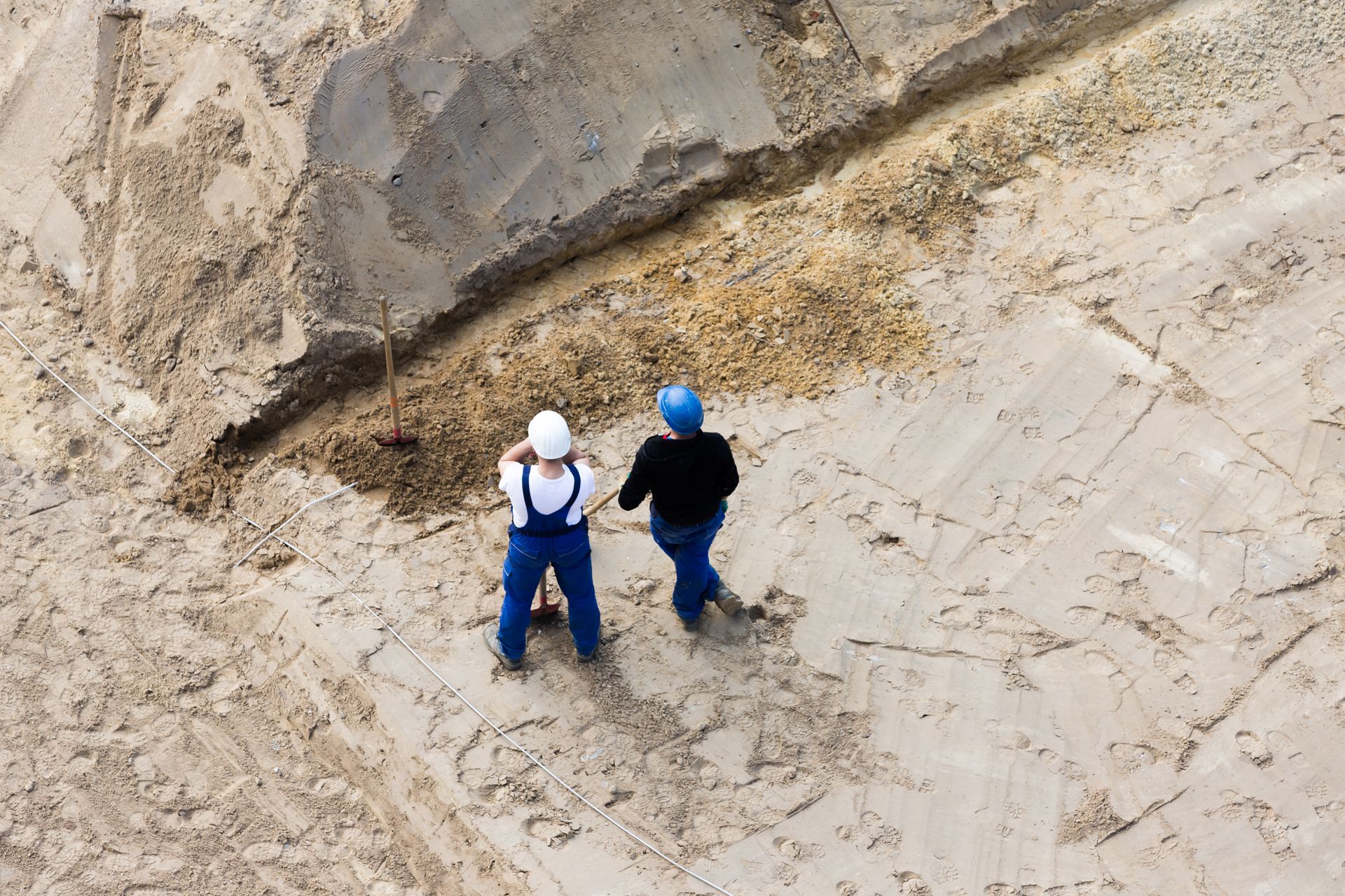 Deux ouvriers, casqués et vêtus de vêtements de travail, se tiennent debout sur un grand chantier sablonneux, à côté d'une tranchée.