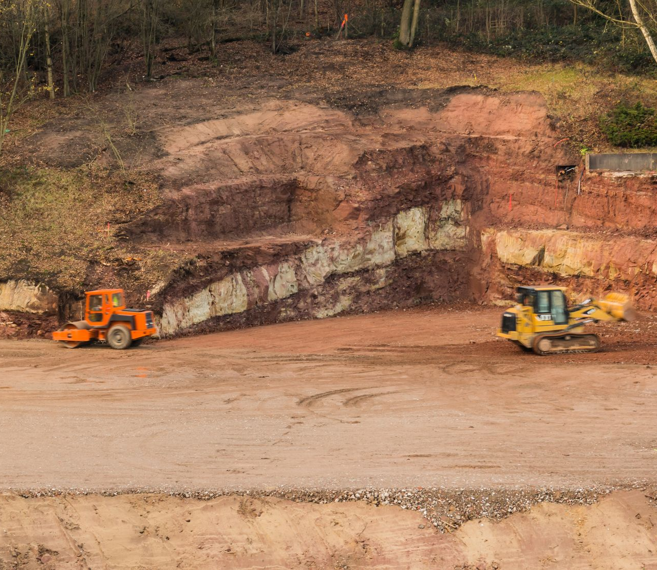 Un chantier de construction avec une excavatrice jaune et un rouleau compresseur orange garés devant une paroi rocheuse excavée et stratifiée.