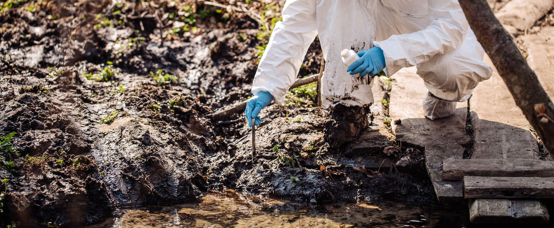 Une personne vêtue d'une combinaison blanche de protection et de gants bleus prélève un échantillon de sol près d'une zone sombre et boueuse.