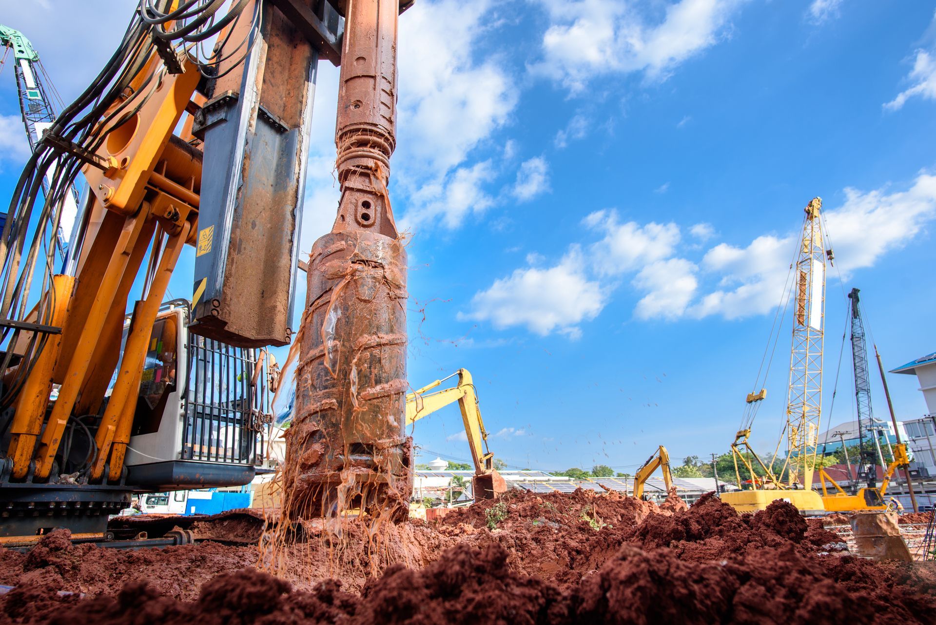 Une imposante foreuse extrait de la terre rouge sur un chantier lumineux et nuageux, d'autres engins étant visibles au loin.