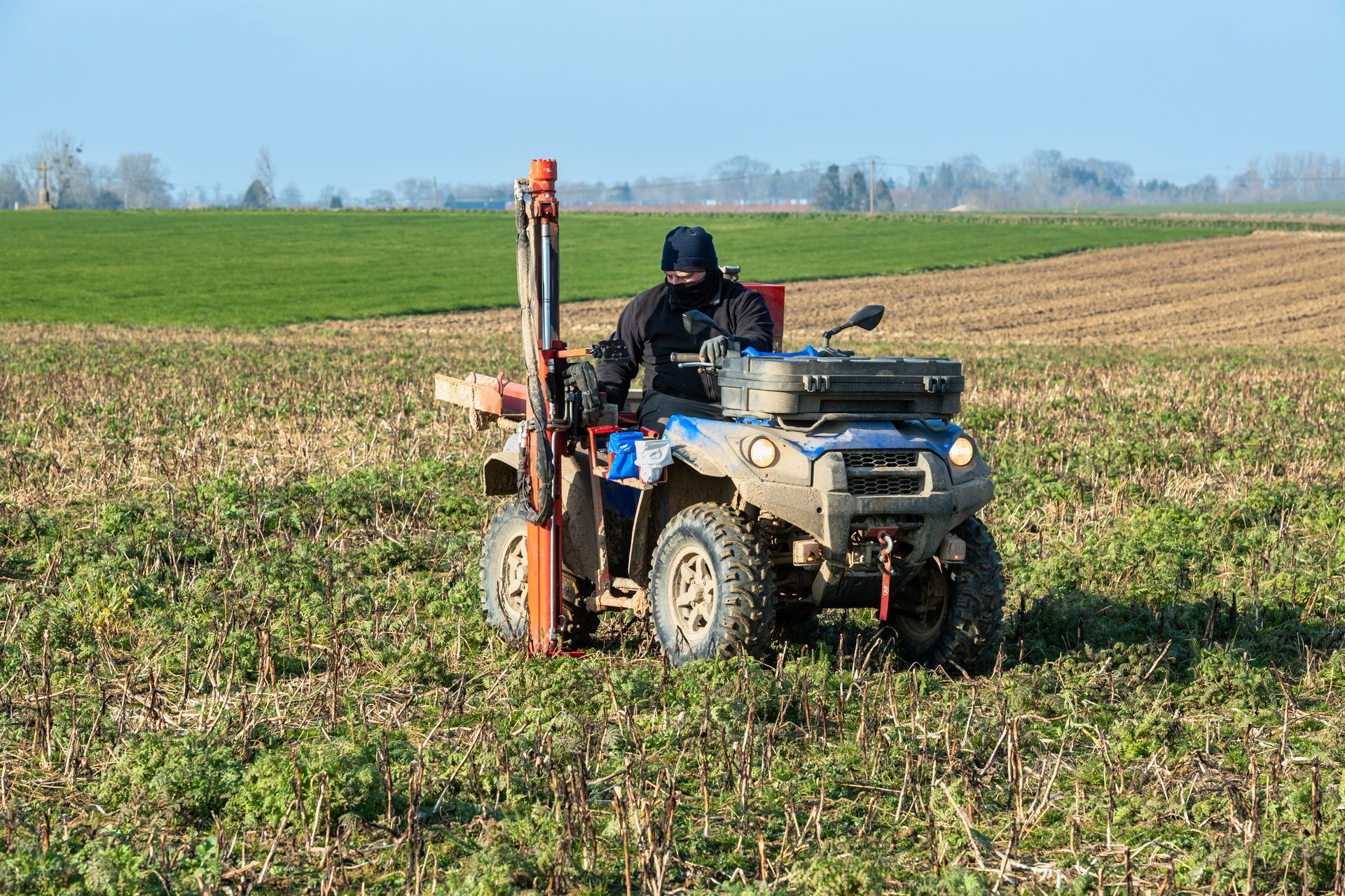 Une personne conduisant un VTT équipé d'une sonde de prélèvement de sol dans un champ, par une journée ensoleillée.