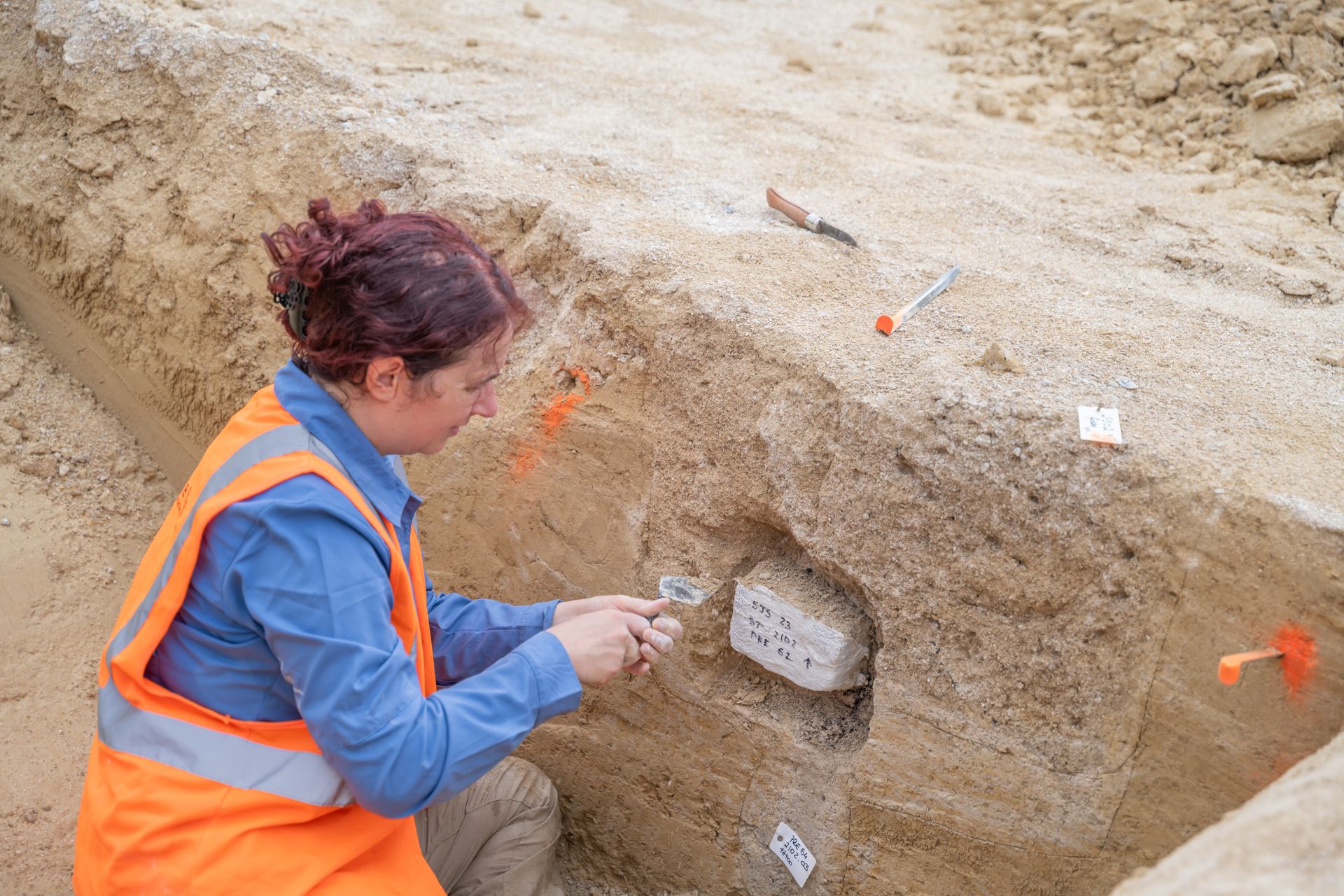 Un archéologue portant un gilet haute visibilité examine un profil de sol dans une tranchée, à l'aide d'un petit outil sur un bloc exposé.