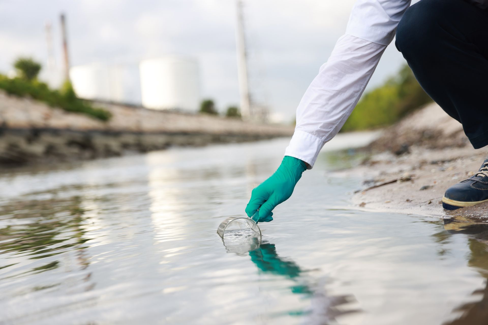 Une personne portant une chemise blanche à manches longues et des gants verts prélève un échantillon d'eau dans un canal situé près de réservoirs industriels.