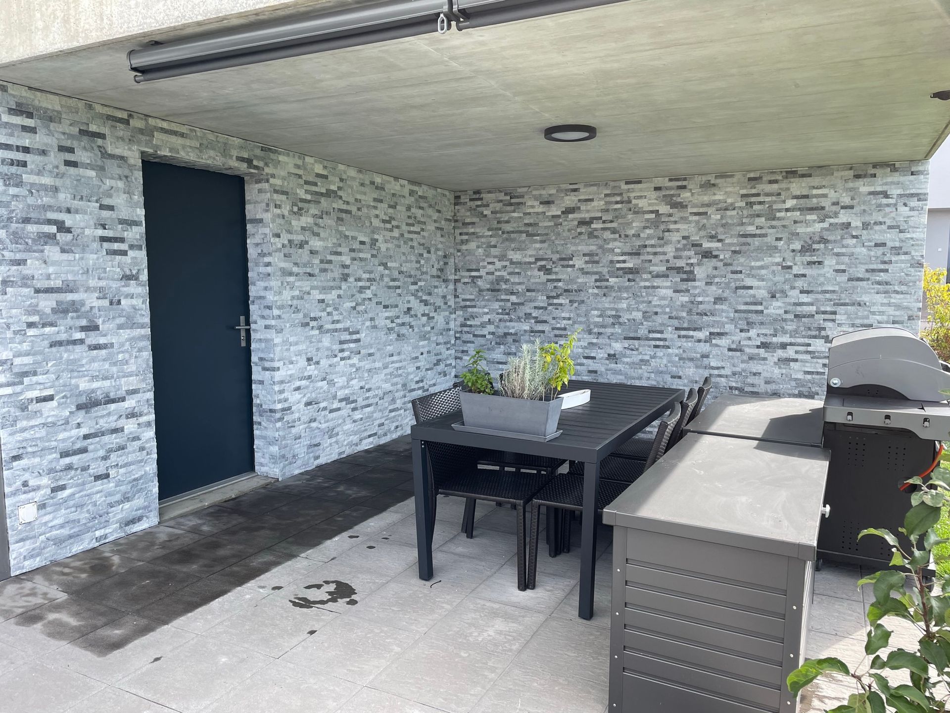 Garage avec mur gris imitation pierre, porte noire et plafond en béton. Sol pavé noir et pelouse verte.