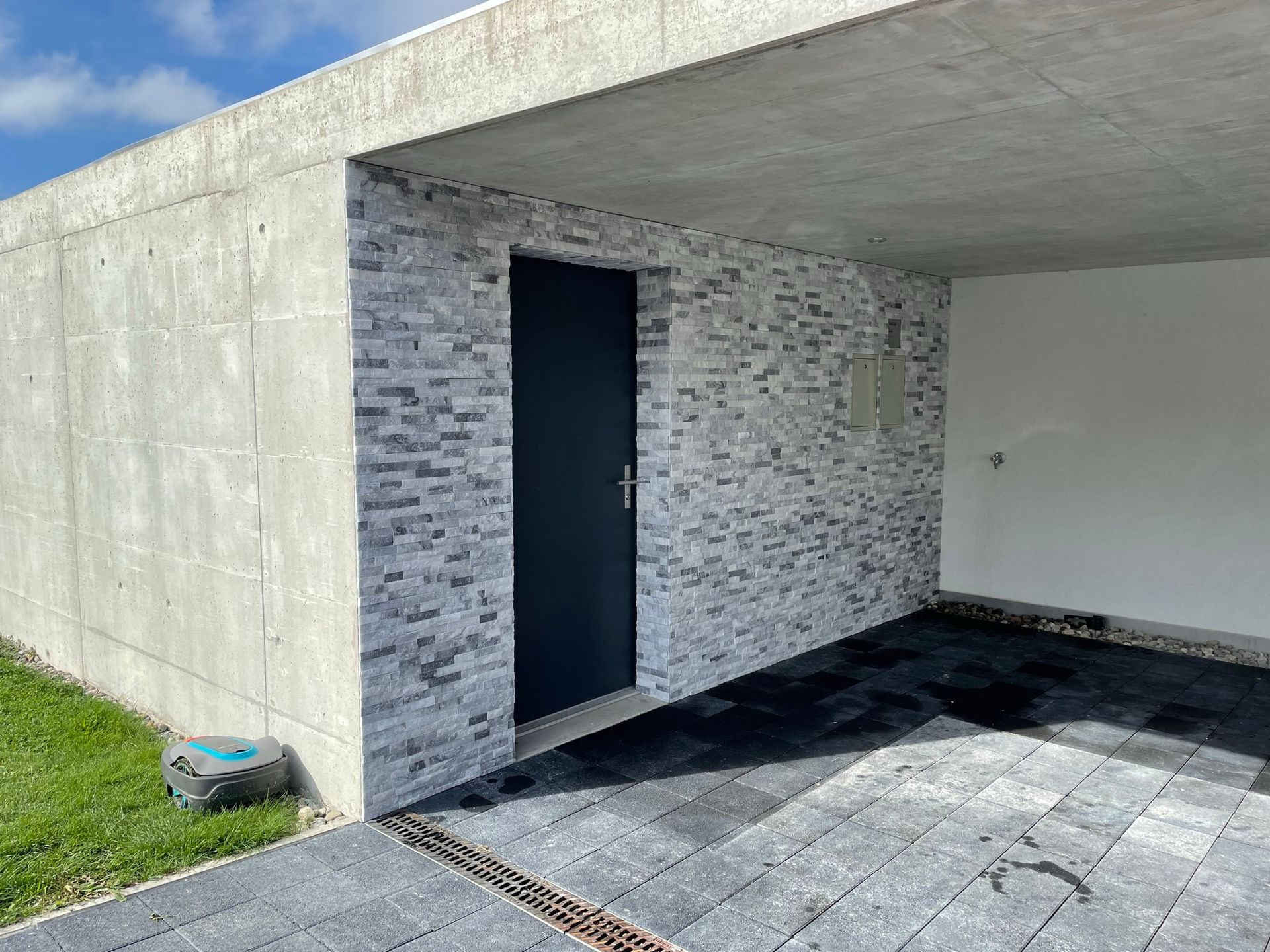 Garage avec mur gris imitation pierre, porte noire et plafond en béton. Sol pavé noir et pelouse verte.