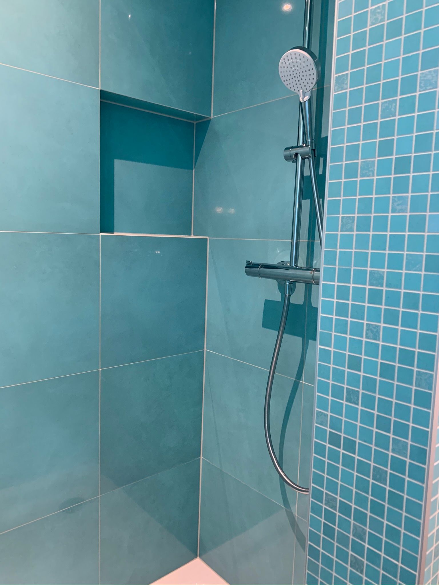 Douche carrelée de bleu avec étagère intégrée, pommeau de douche et mur recouvert de petits carreaux carrés.