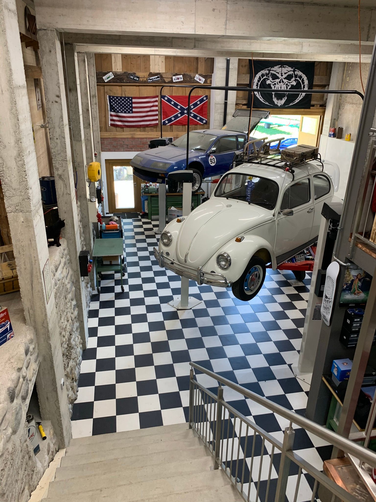Une Coccinelle Volkswagen blanche sur un pont élévateur dans un garage au sol à damier, avec des drapeaux américain et confédéré au mur.