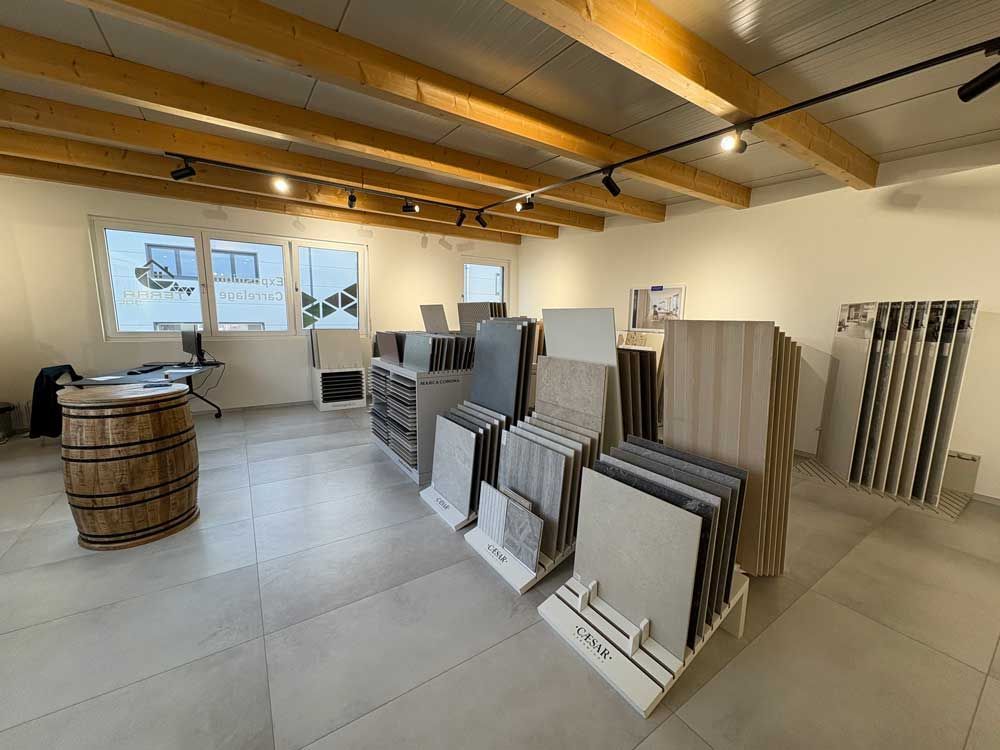Intérieur d'une salle d'exposition de carrelage avec des échantillons présentés. On aperçoit un tonneau en bois, des fenêtres et des rails d'éclairage.