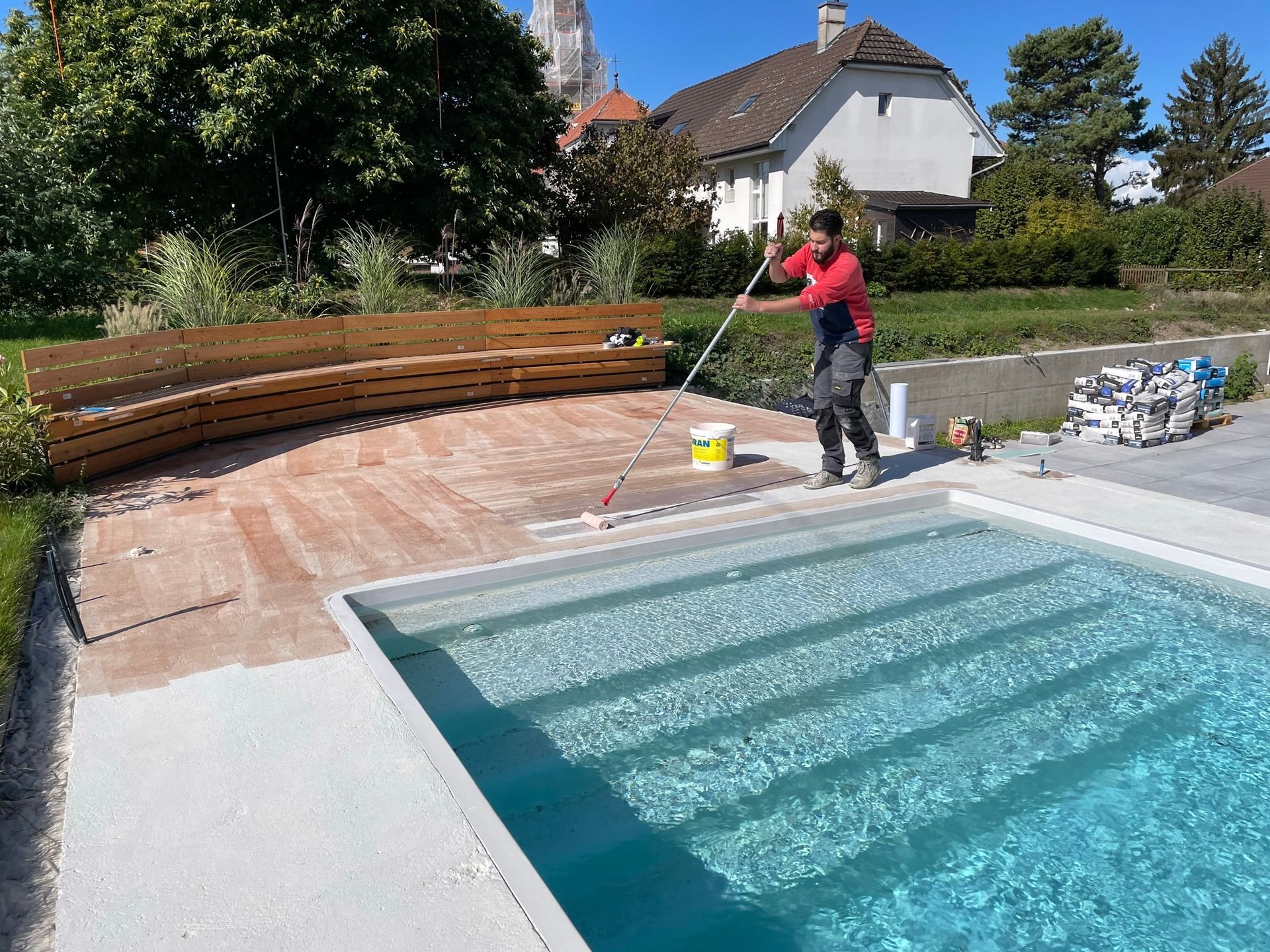 Un homme travaille sur la terrasse d'une piscine. Un banc en bois incurvé se trouve à proximité, et la piscine est remplie d'eau.