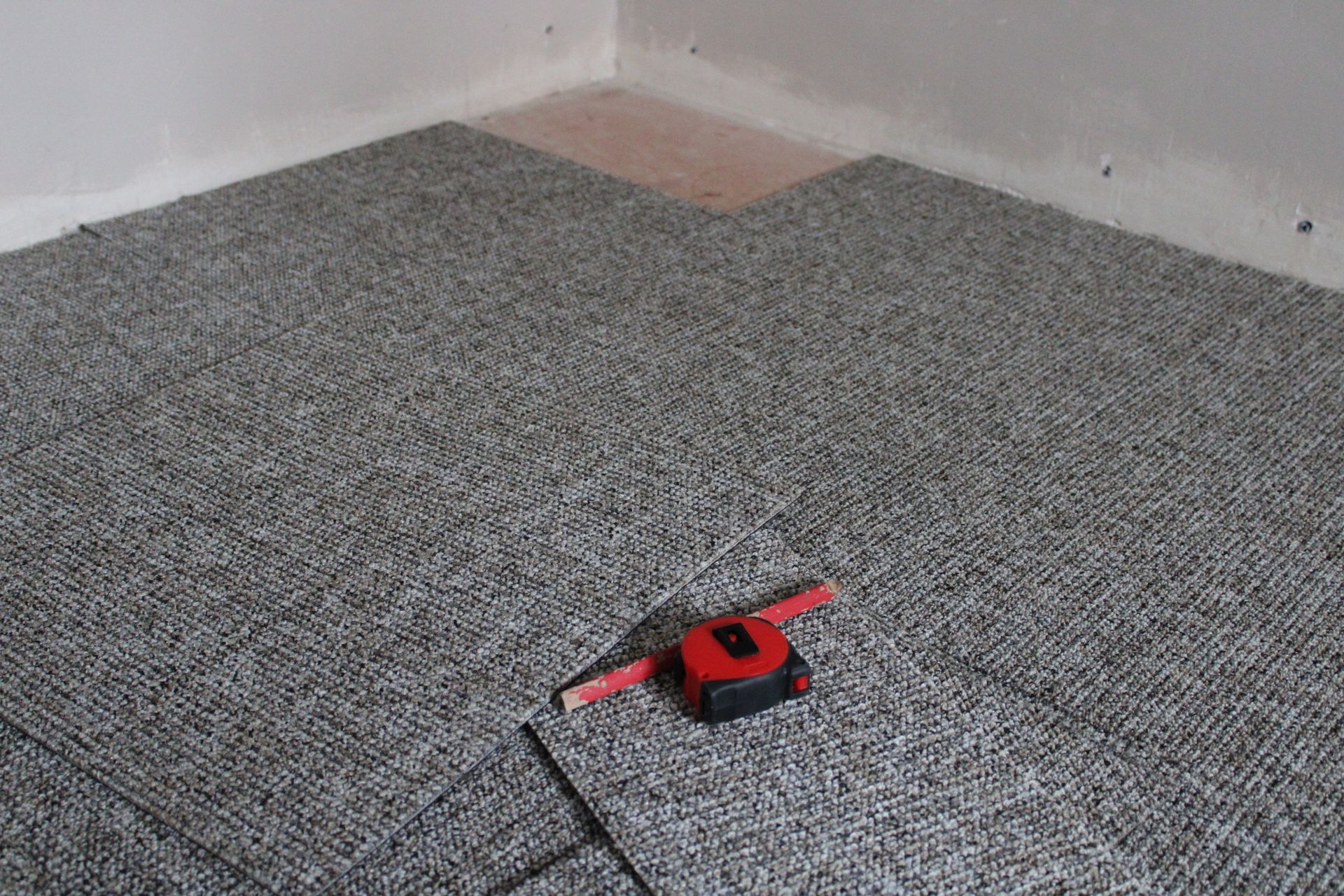 Pose de dalles de moquette en cours ; le sol est recouvert de dalles grises. Un ruban métrique rouge est apposé sur la surface près de l'angle.