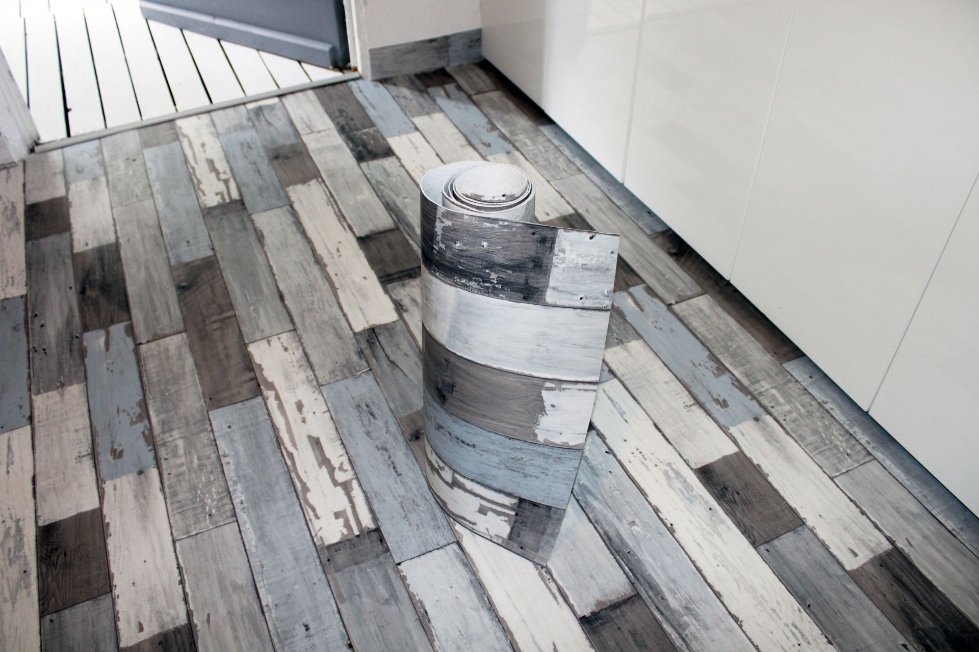 Échantillon de revêtement de sol en vinyle enroulé, présentant un motif de lames de bois dans des tons gris et bleus sur un plancher en bois.