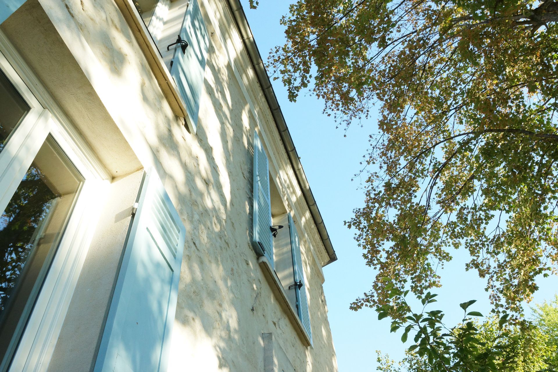Bâtiment couleur crème avec des volets bleu clair, des branches d'arbres et un ciel bleu clair.
