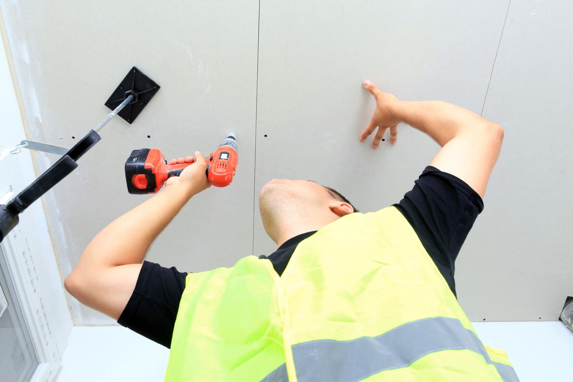 Personne installant des plaques de plâtre au plafond à l'aide d'une perceuse ; portant un gilet de sécurité jaune.