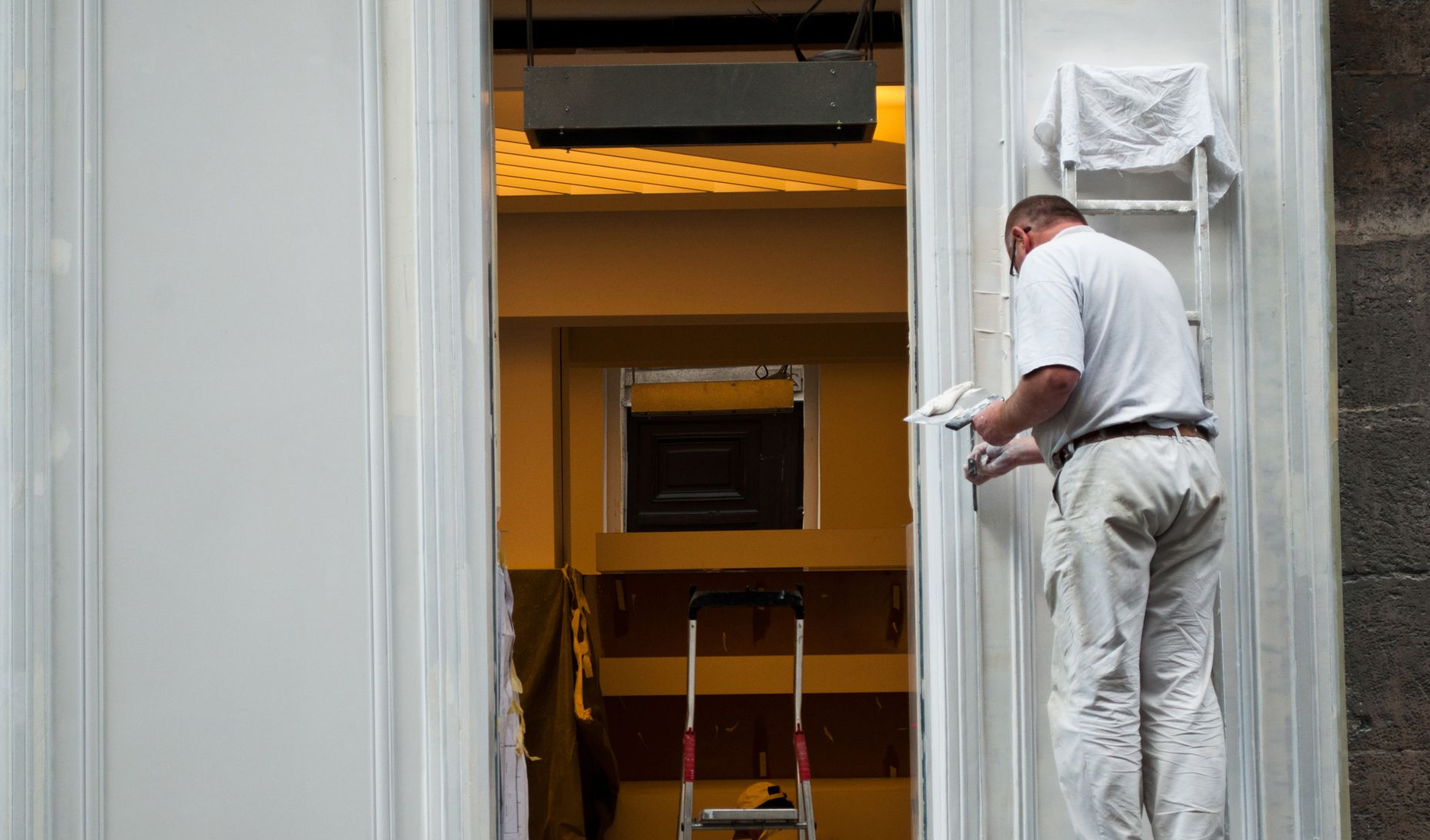Un peintre en chemise et pantalon blancs peint un encadrement de porte blanc.