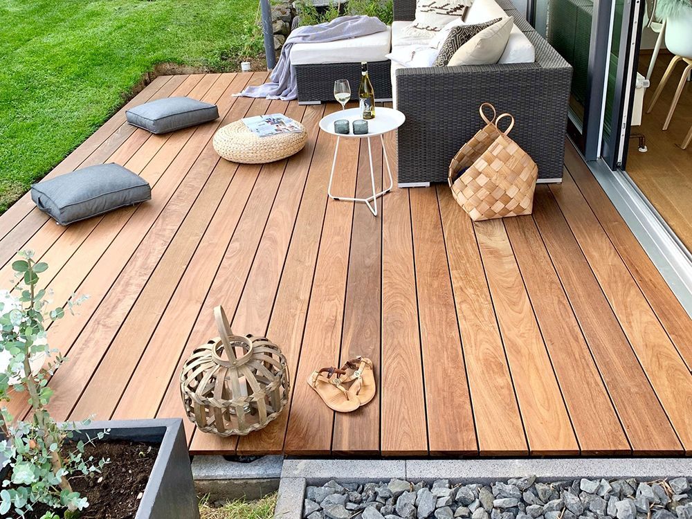 Holzterrasse mit Sitzgruppe, Zierkissen und kleinem Tisch. Angrenzend an eine grüne Rasenfläche und eine Glasschiebetür.