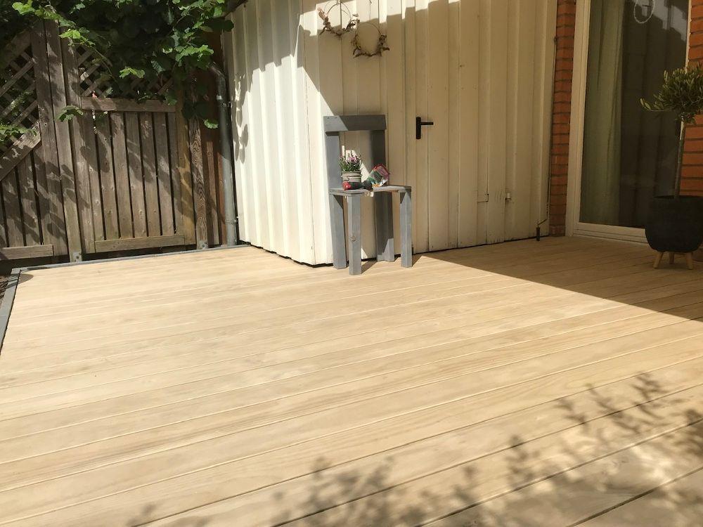 Eine Holzterrasse mit einem kleinen Tisch vor einem weißen Schuppen, einem Holzzaun und einer Topfpflanze.