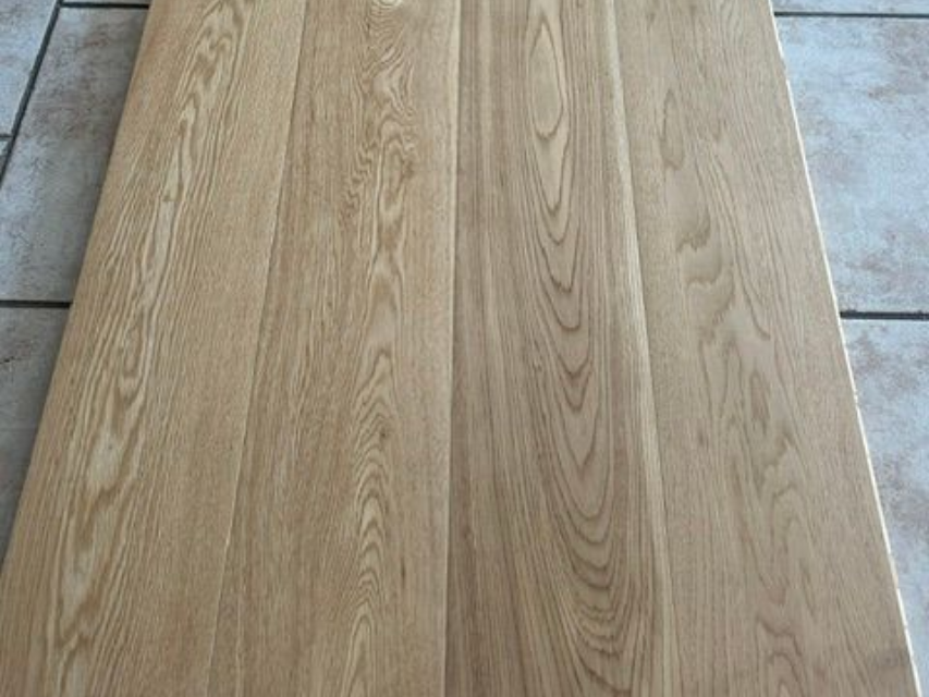 Eine helle Holztischplatte mit sichtbarer Maserung steht mittig auf einem gefliesten Boden.