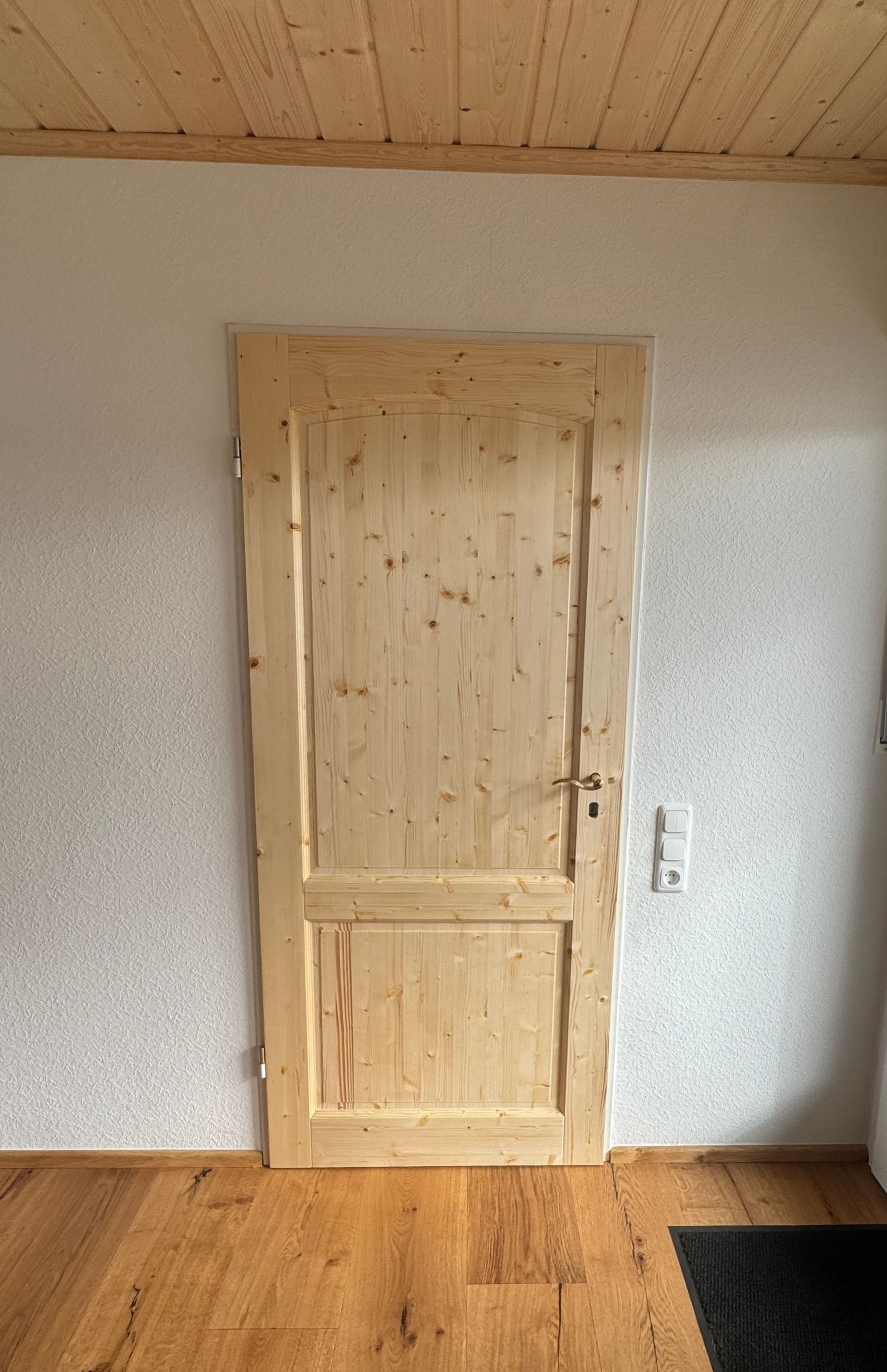 Eine helle Holztür mit zweiflügeligem Design in einem Raum mit weiß strukturierten Wänden und Holzboden.