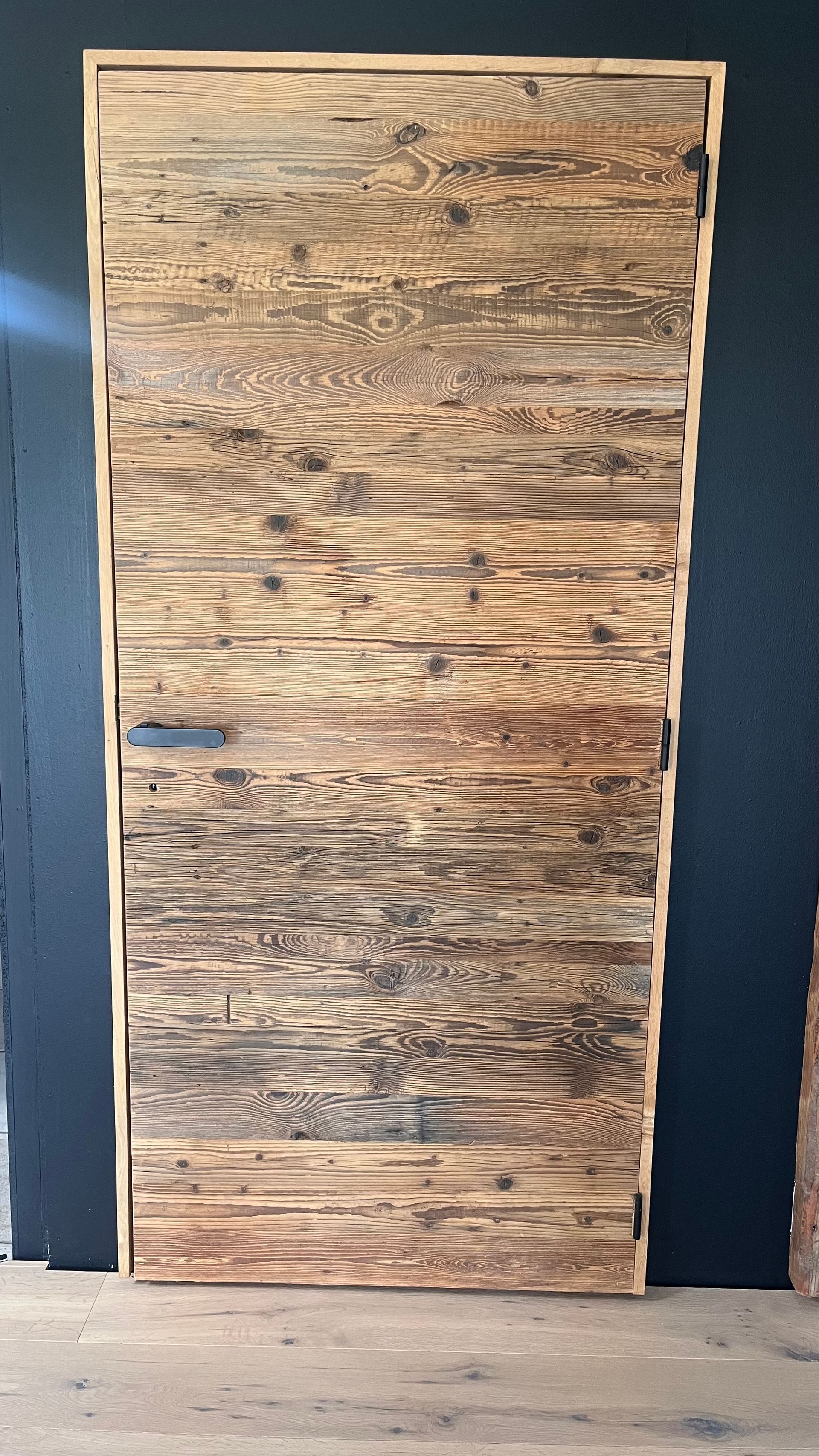 Eine rustikale, hellbraune Holztür mit horizontaler Maserung, eingerahmt von einer dunklen, einfarbigen Wand.