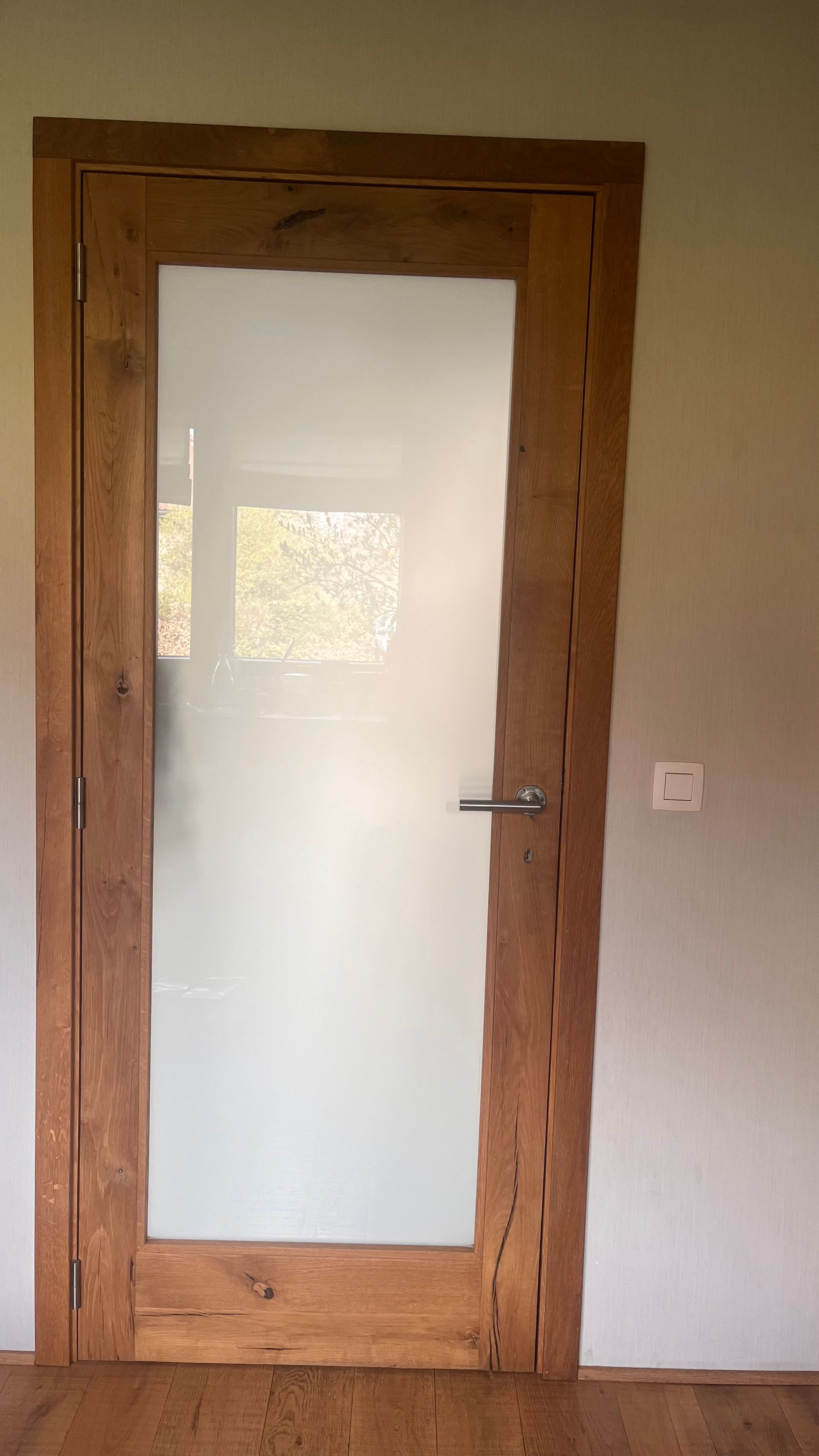 Eine rustikale Holztür mit einer großen Milchglasscheibe und einem Metallgriff, eingefasst in eine schlichte, helle Wand.