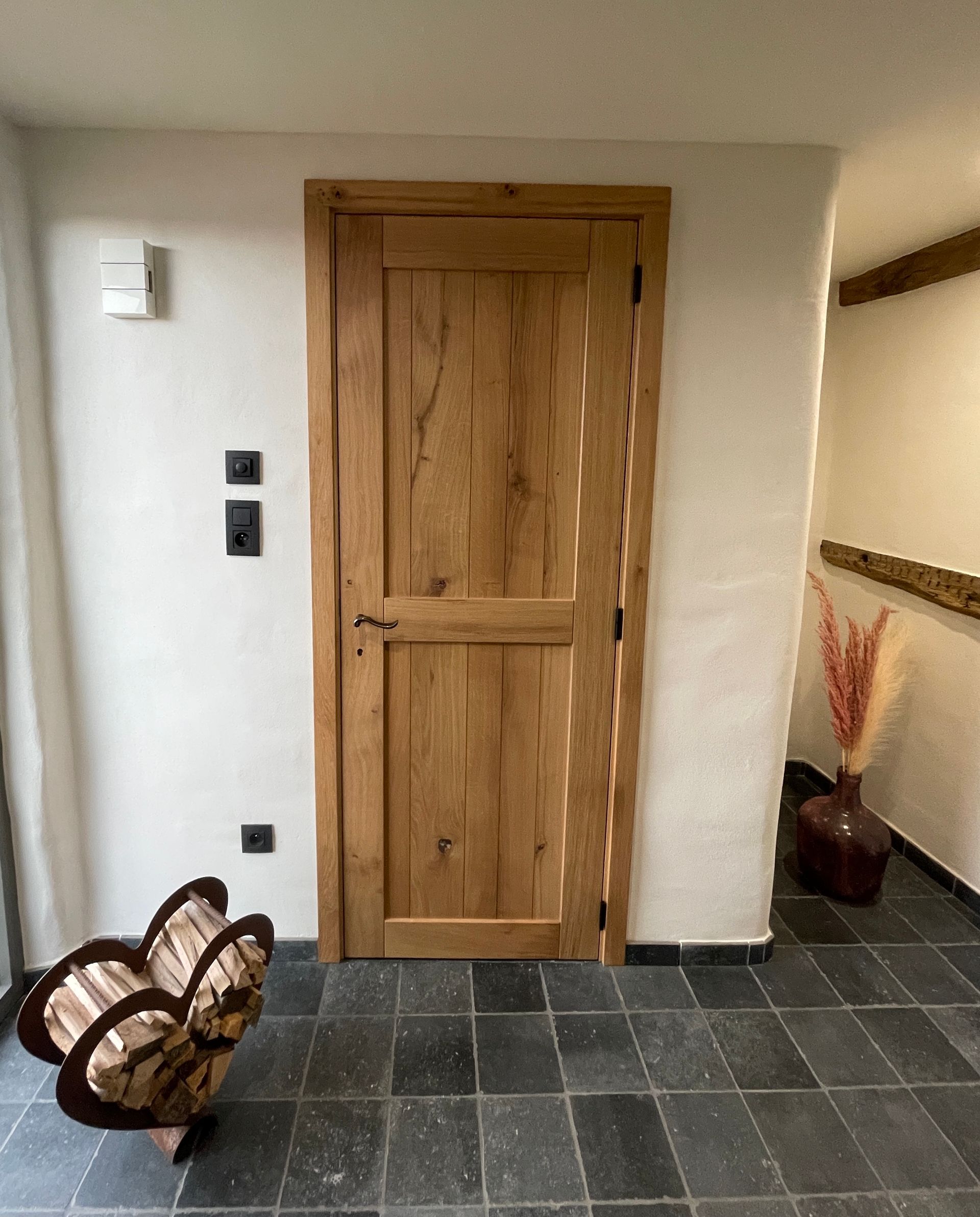 Eine rustikale Holztür vor einer weißen Wand, dazu ein dunkler Fliesenboden, ein dekorativer Holzständer und eine Vase mit Trockenblumen.