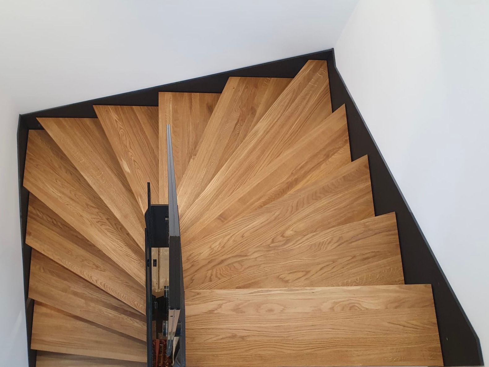 Draufsicht auf eine Wendeltreppe mit hellen Holzstufen und einem schwarzen Mittelgeländer vor weißen Wänden.