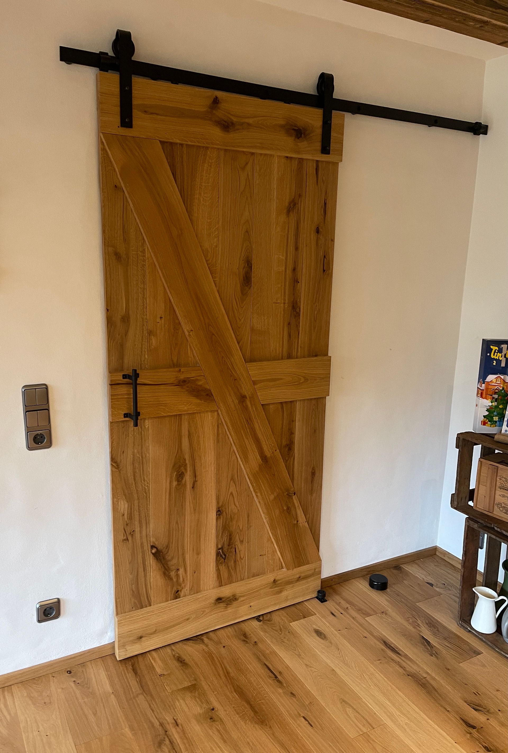 Eine rustikale, helle Holzschiebetür im Scheunenstil mit schwarzem Eisengriff und Beschlägen, die an einer Wand in einem Zimmer mit Holzboden montiert ist.