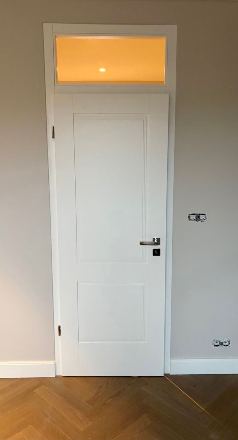 Eine geschlossene weiße Innentür mit einem rechteckigen Oberlicht darüber, eingelassen in eine beige Wand mit Fischgrätparkett.