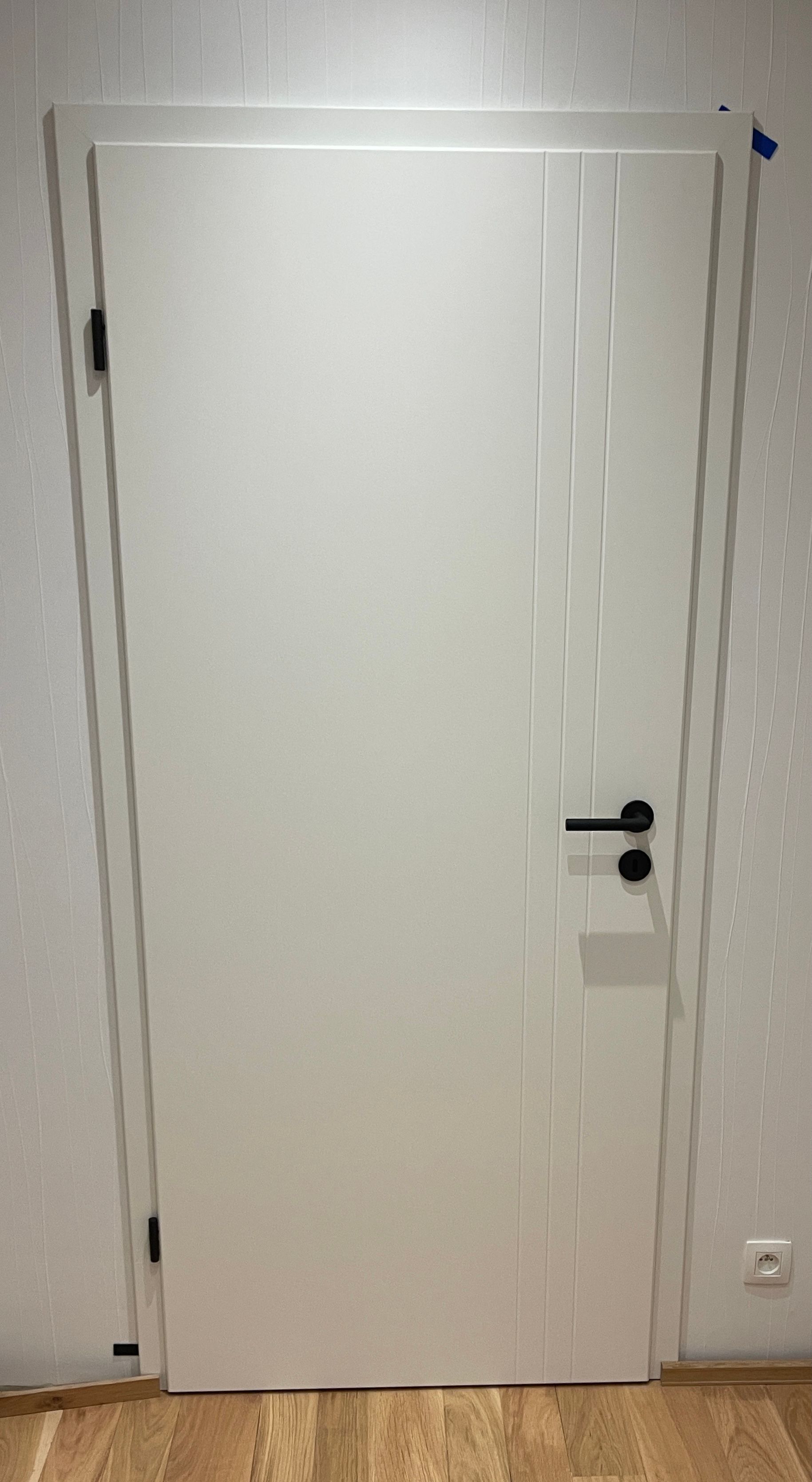 Eine weiße Innentür mit vertikalen Zierrillen, schwarzem Griff und Scharnieren, eingebaut in einem hellen Raum.