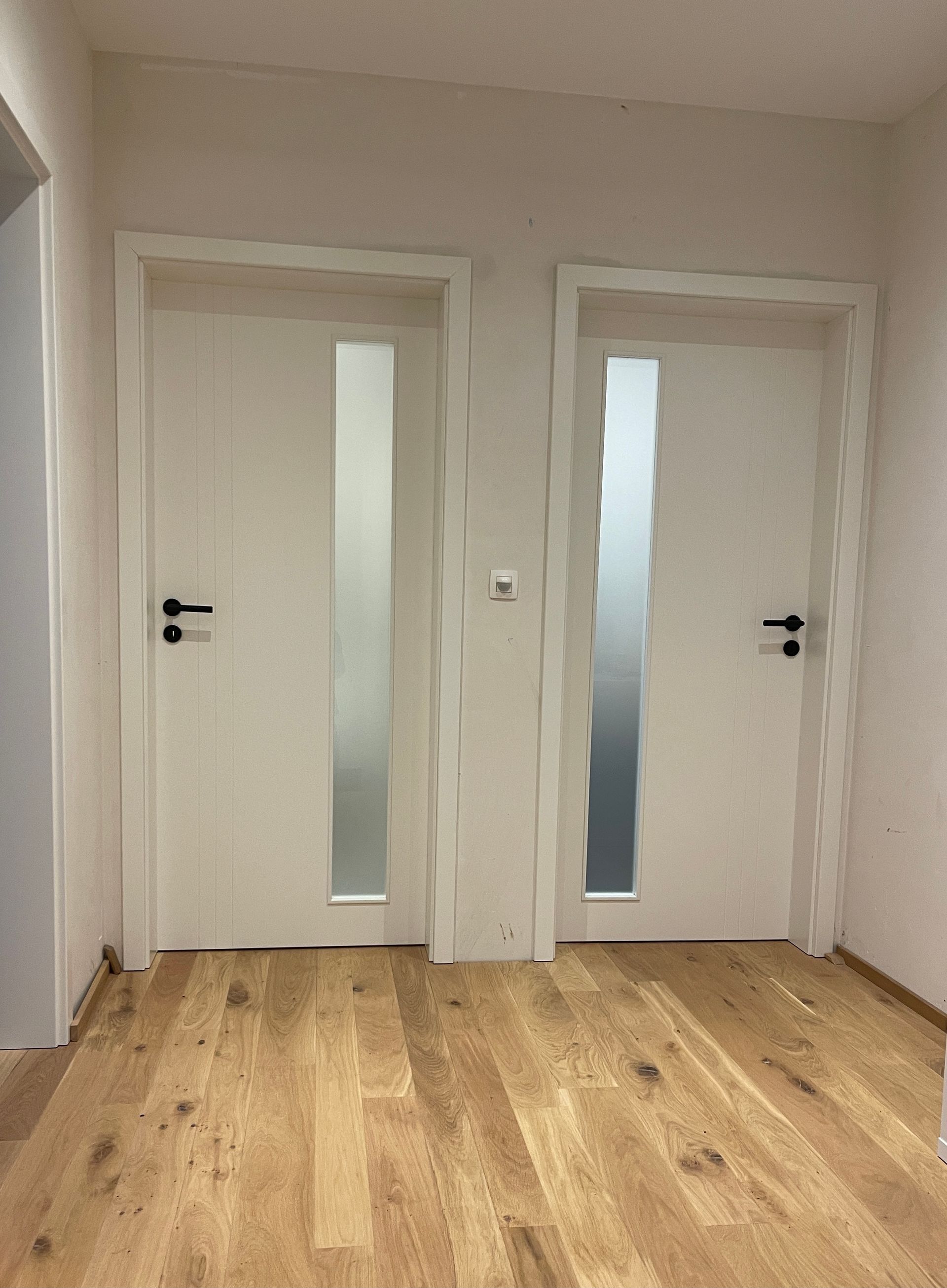 Zwei weiße Innentüren mit vertikalen Milchglasscheiben und schwarzen Griffen in einem Flur mit Holzboden.