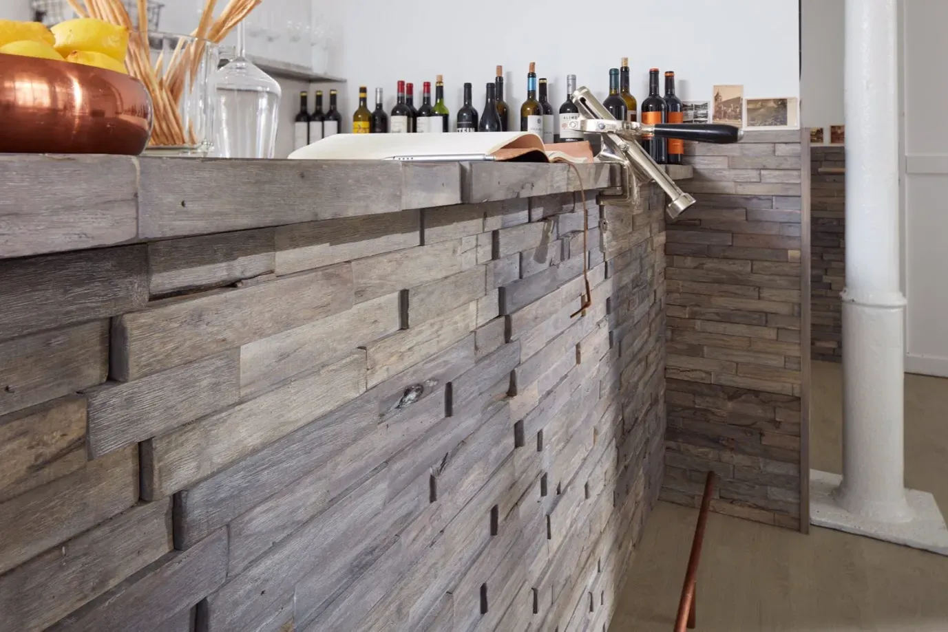 Eine hölzerne Bar mit Flaschen und einer Kupferschale; im Hintergrund eine weiße Säule.