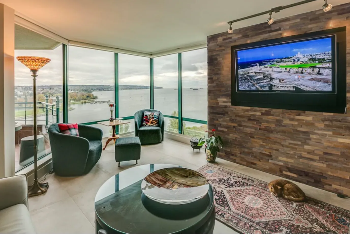 Wohnzimmer mit Meerblick, dunkelgrünen Stühlen, rundem Couchtisch, an der Backsteinwand montiertem Fernseher und Teppich.