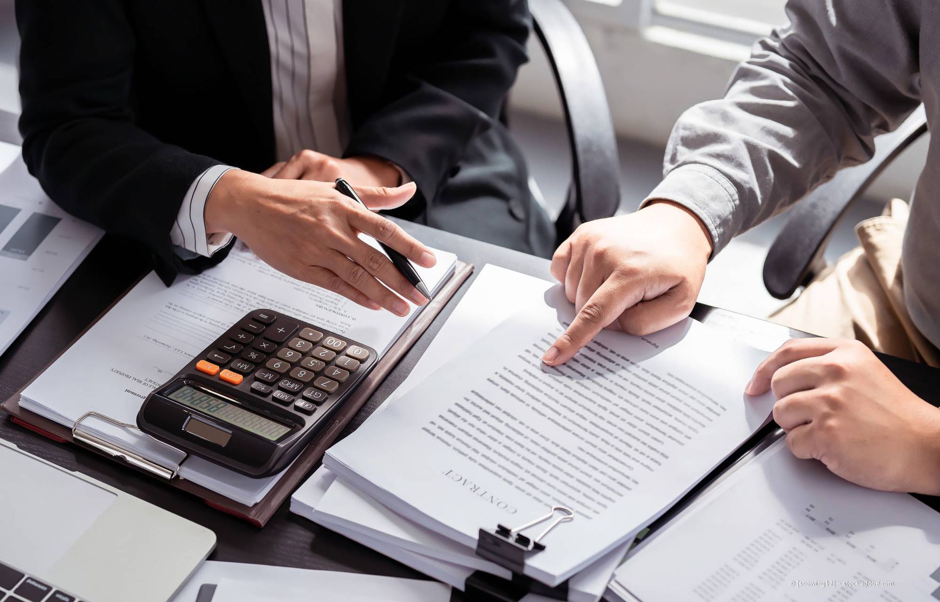 Zwei Personen prüfen Dokumente mit einem Taschenrechner. Eine Person zeigt auf den Text, die andere gestikuliert.