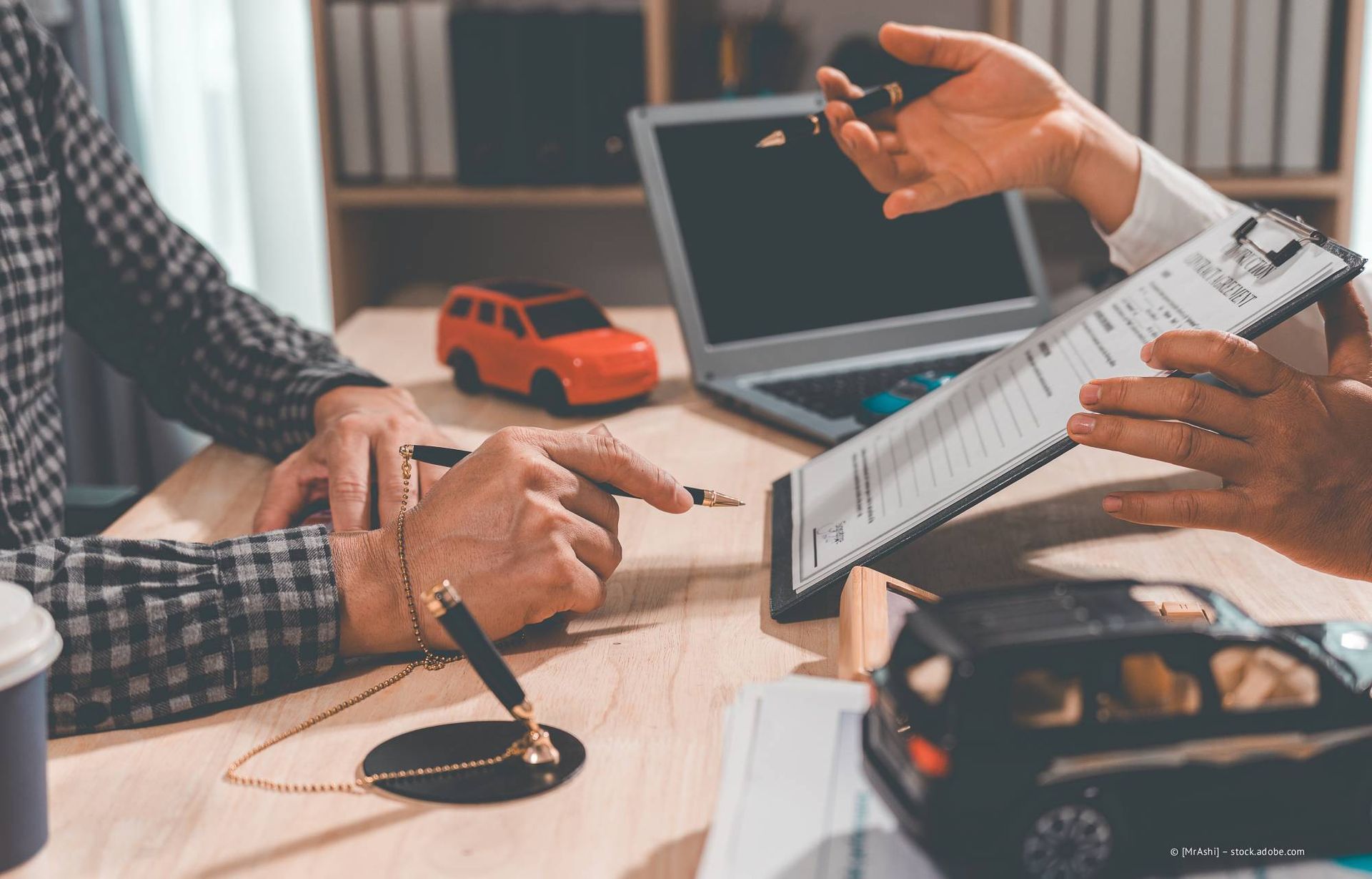 Eine Person unterzeichnet den Autokaufvertrag, eine andere Person hält die Autoschlüssel, das Automodell steht auf dem Schreibtisch.